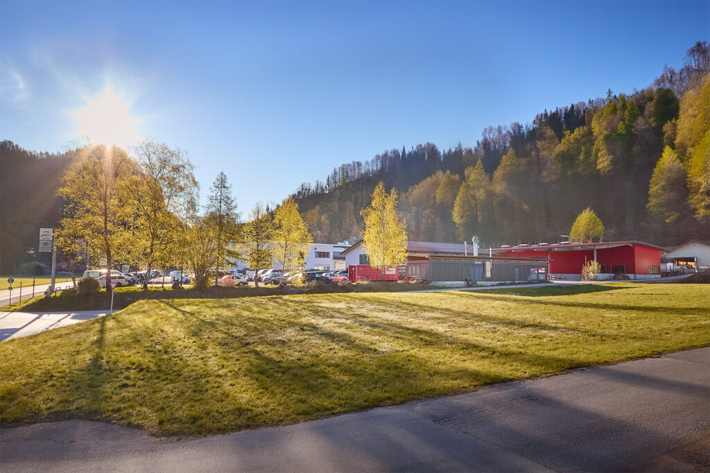 Ein moderner Gebäudekomplex mit roten und weißen Strukturen steht auf einer sonnenbeschienenen Wiese, umgeben von geparkten Autos, Bäumen und einem bewaldeten Hügel im Hintergrund. LAC Berger Industrielackierung