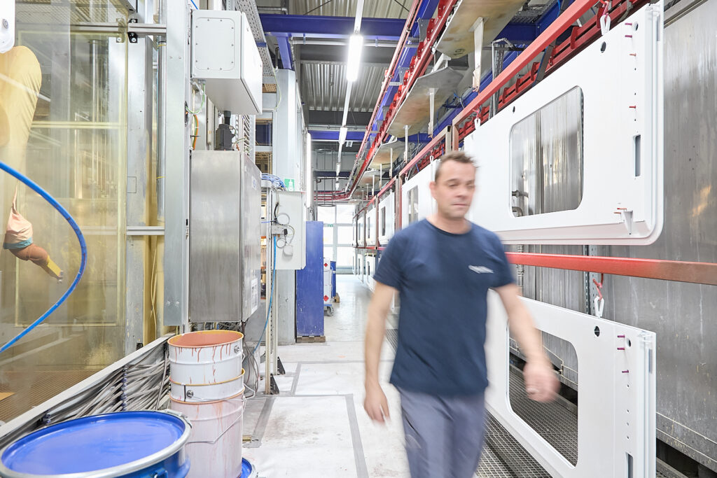 Eine Person in einem blauen Hemd geht durch eine hell erleuchtete Industrieanlage, in der weiße Platten, Metallgeräte und Farbeimer zu sehen sind. LAC Berger Industrielackierung
