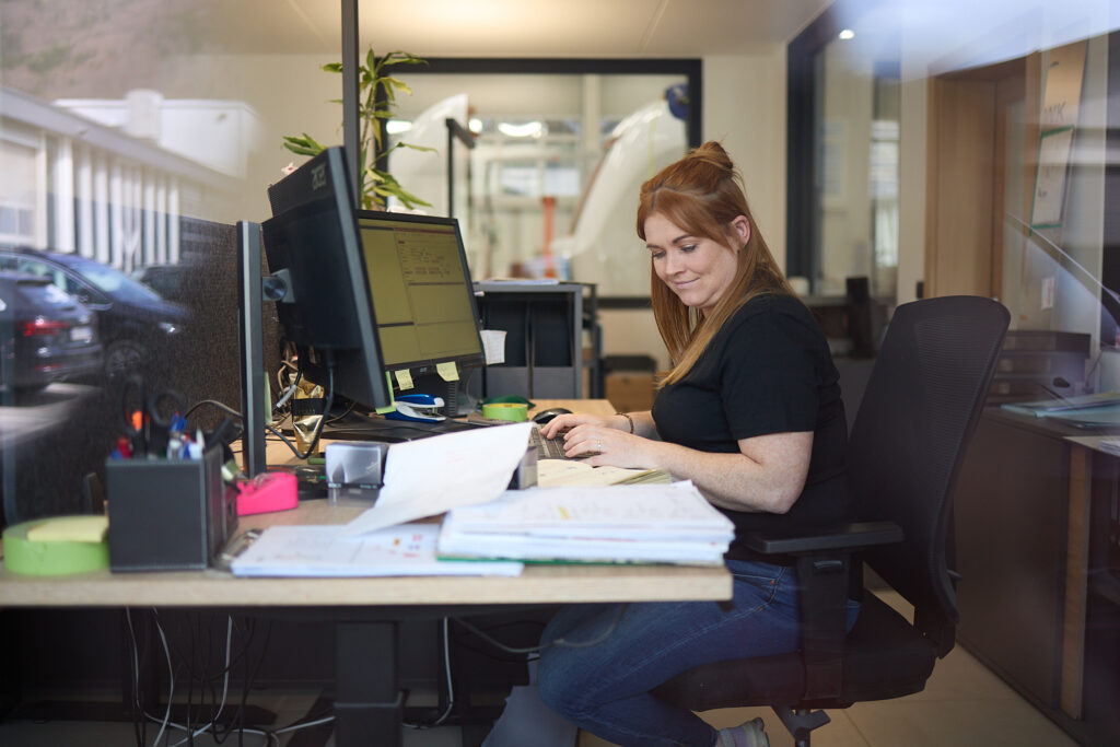 Eine Frau sitzt an einem Schreibtisch in einem Büro und arbeitet an einem Computer, während um sie herum Papierkram und Büromaterialien verstreut sind. LAC Berger Industrielackierung