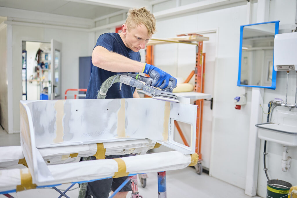 Eine Person mit Handschuhen bearbeitet ein grundiertes Autoteil in einer sauberen, gut beleuchteten Werkstatt mit einem Elektroschleifer. LAC Berger Industrielackierung