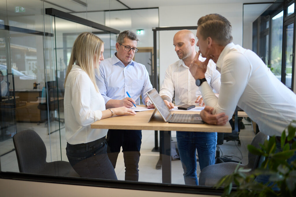 Vier Personen stehen um einen Stehtisch in einem Büro und diskutieren während einer Besprechung über Dokumente und einen Laptop. LAC Berger Industrielackierung
