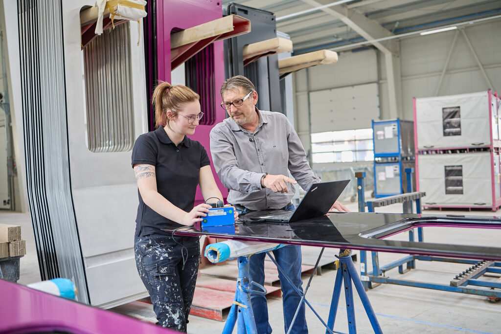 Zwei Personen in einer Fabrikumgebung schauen auf einen Laptop auf einem Arbeitstisch, im Hintergrund sind große lackierte Metallteile und Industrieanlagen zu sehen. LAC Berger Industrielackierung