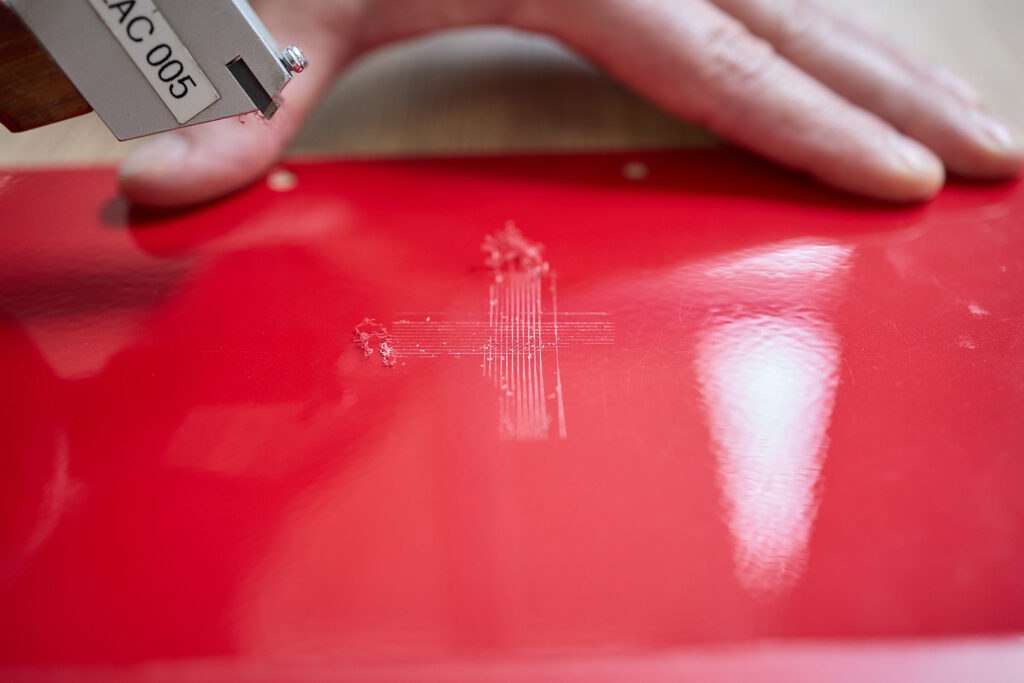 Eine Hand hält eine rote Oberfläche, auf der mit einem speziellen Gerät ein Kreuzkratztest zur Beurteilung der Beschichtungshaftung durchgeführt wird. LAC Berger Industrielackierung