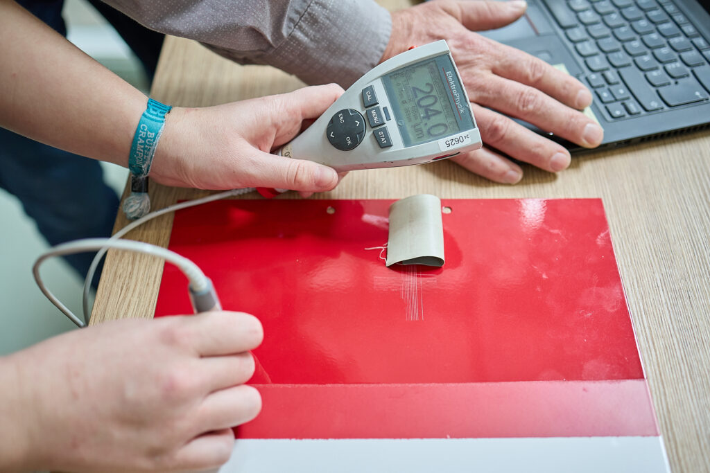 Eine Person benutzt ein digitales Dickenmessgerät auf einer rot-weißen Fläche, während eine andere Hand neben einer Laptop-Tastatur ruht. LAC Berger Industrielackierung