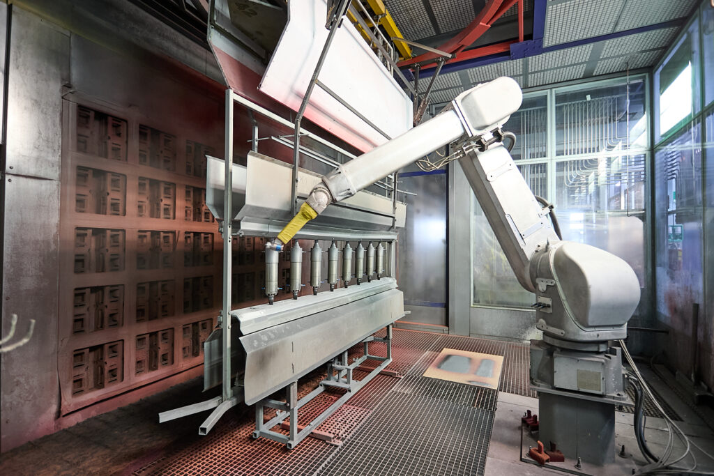 Ein Industrieroboterarm arbeitet in einer automatischen Lackierkabine und beschichtet Metallteile, die für die Produktion aufgereiht sind. LAC Berger Industrielackierung
