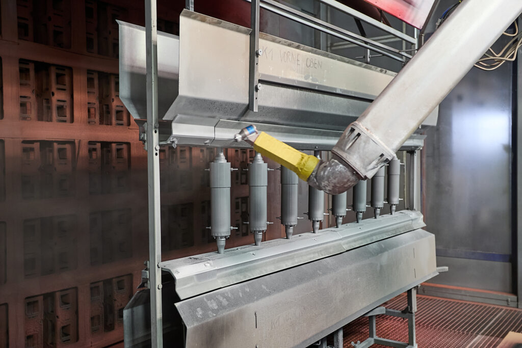 Ein Roboterarm trägt die Pulverbeschichtung auf zylindrische Metallteile auf, die in einer industriellen Umgebung auf einem Gestell angeordnet sind. LAC Berger Industrielackierung