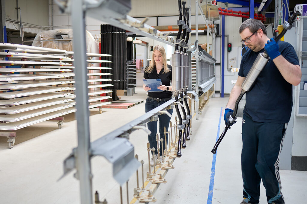 Eine Frau prüft ein Klemmbrett, während ein Mann in einer Fabrikhalle mit Metallteilen arbeitet, die auf Regalen im Hintergrund aufgereiht sind. LAC Berger Industrielackierung