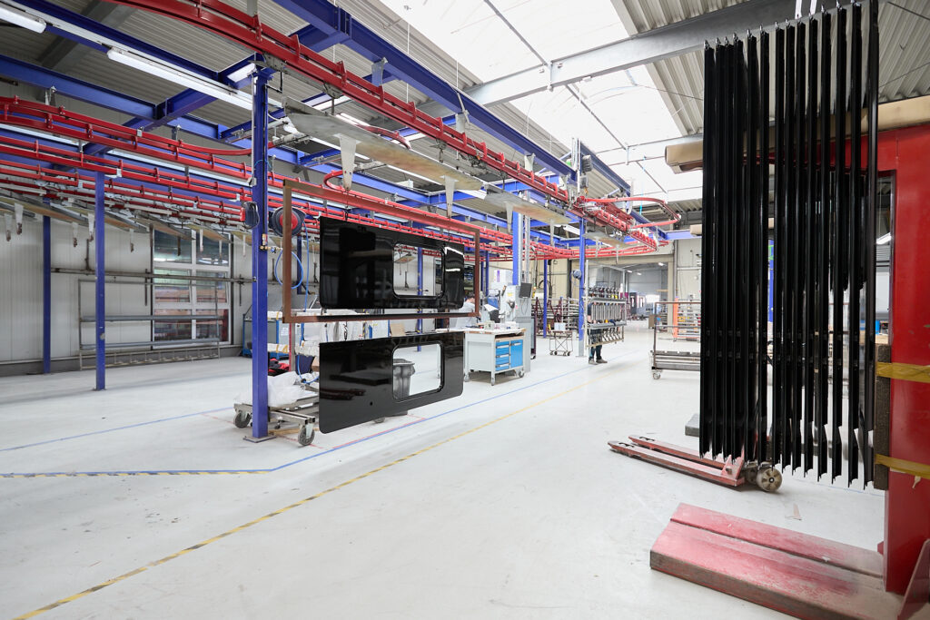 Große, saubere Industriehalle mit Metallkonstruktionen, aufgehängten schwarzen Tafeln, Arbeitstischen und Hängebahnsystem; helle Beleuchtung durch Oberlichter. LAC Berger Industrielackierung