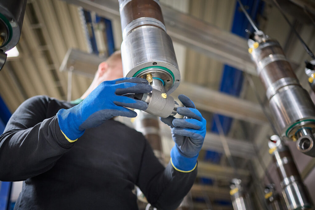 Eine Person mit blauen Handschuhen prüft ein Metallteil in einer industriellen Umgebung mit Maschinen im Hintergrund. LAC Berger Industrielackierung