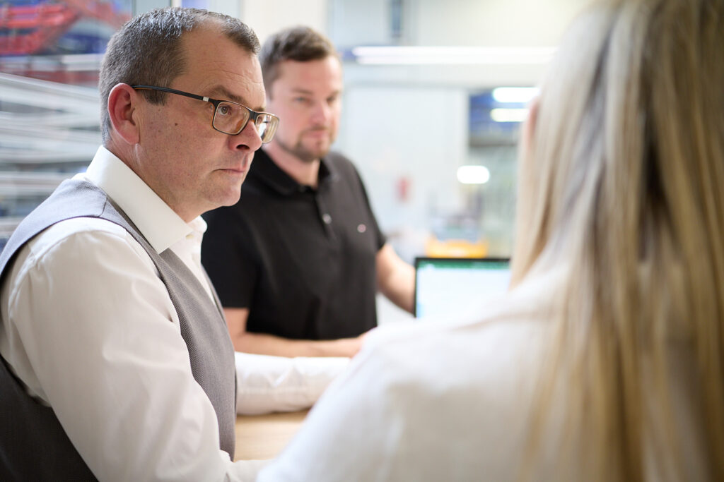 Drei Personen führen ein Gespräch in einem Büro; ein Mann mit Brille ist im Fokus, ein anderer Mann und eine Frau sind im Hintergrund unscharf. LAC Berger Industrielackierung