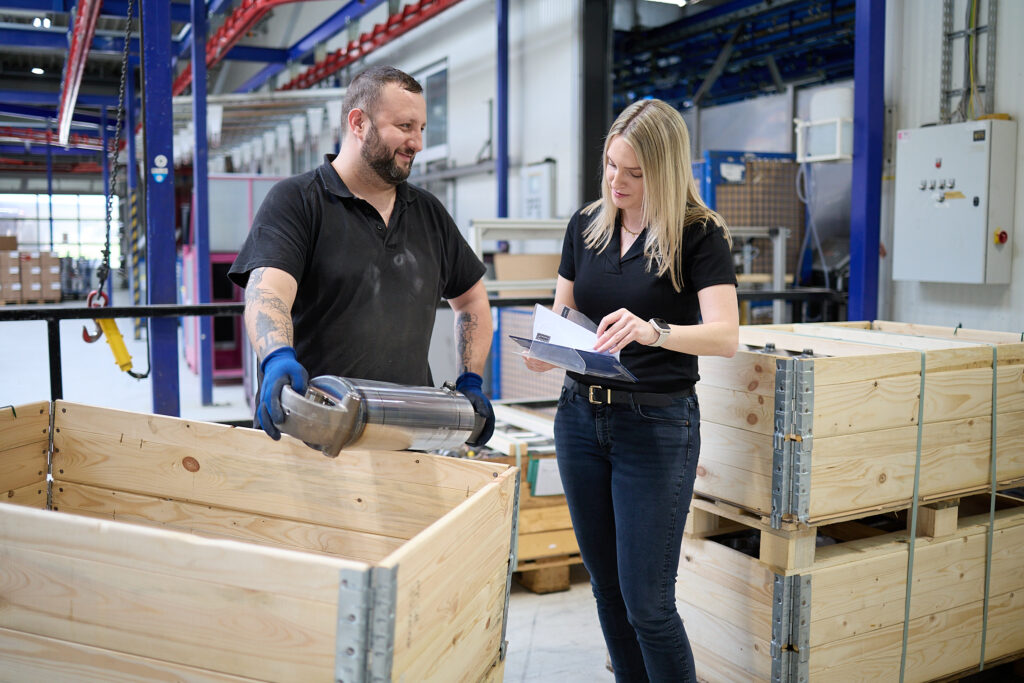 Ein Mann mit einem Metallzylinder in der Hand spricht mit einer Frau, die in einem Lagerhaus neben einer Holzkiste steht und Papiere in der Hand hält. LAC Berger Industrielackierung