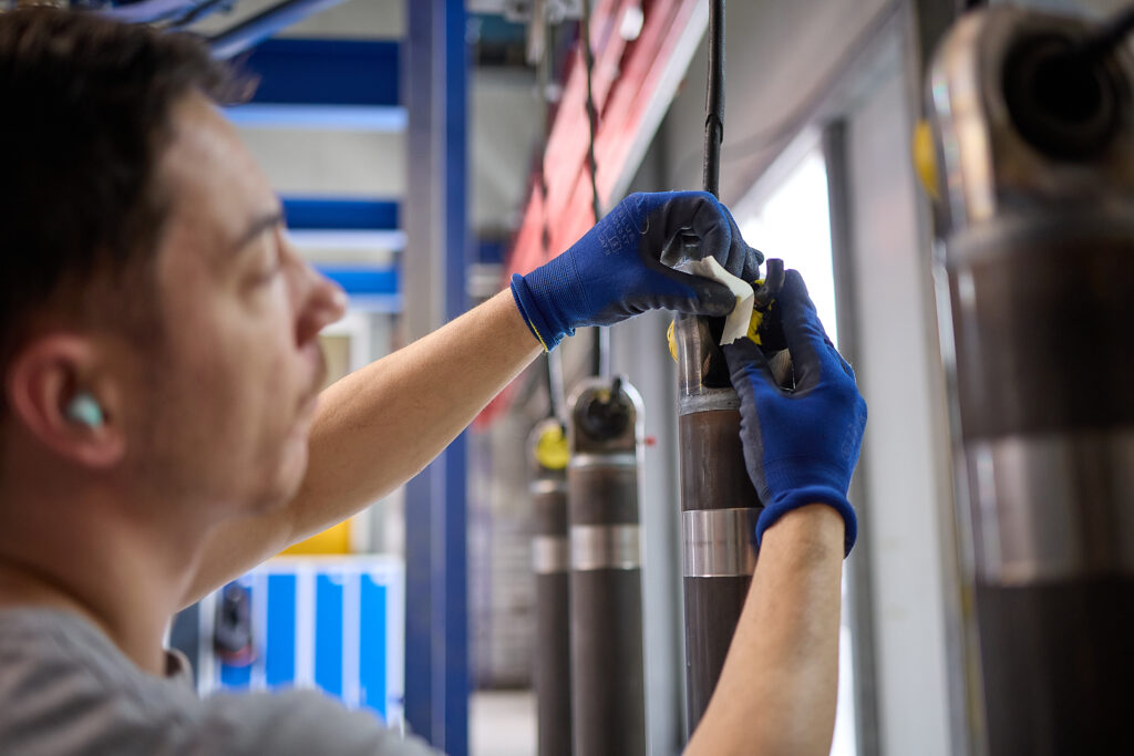 Eine Person mit blauen Handschuhen repariert oder prüft einen Metallzylinder in einer industriellen Umgebung, wobei sie ein kleines Werkzeug oder Material an dem Bauteil verwendet. LAC Berger Industrielackierung