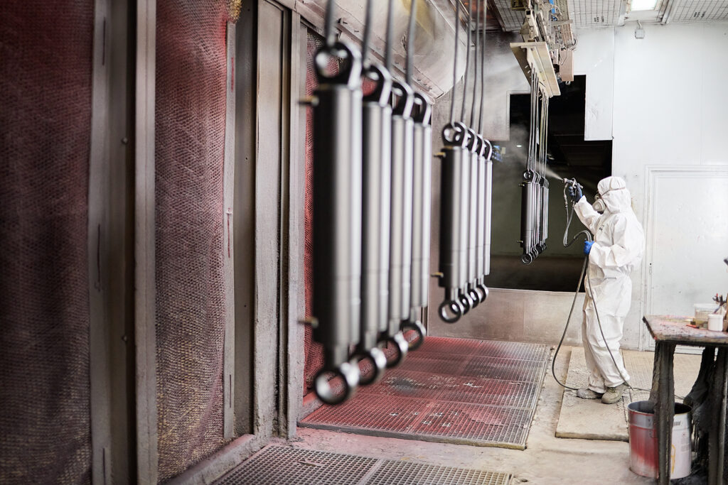 Eine Person in Schutzkleidung beschichtet mit einer Spritzpistole Metallteile, die an einem Fördersystem in einer industriellen Lackieranlage hängen. LAC Berger Industrielackierung
