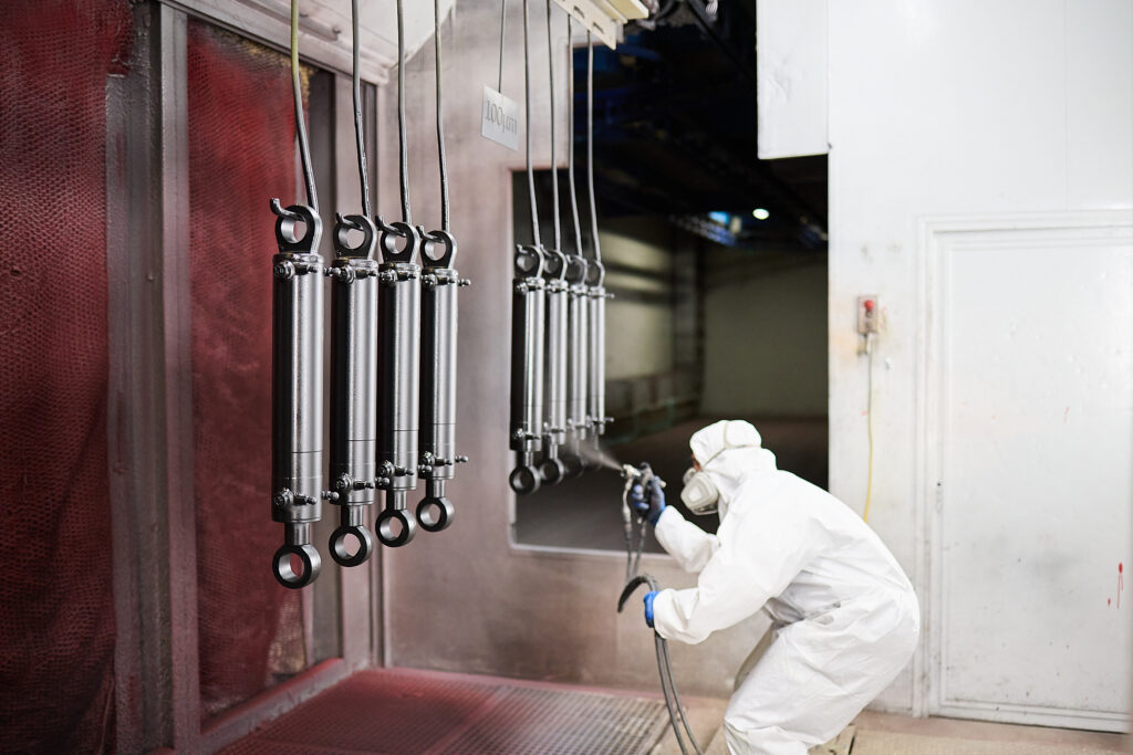 Ein Arbeiter in Schutzkleidung lackiert Metallzylinder, die in einer Reihe in einer Industrieanlage aufgehängt sind. LAC Berger Industrielackierung