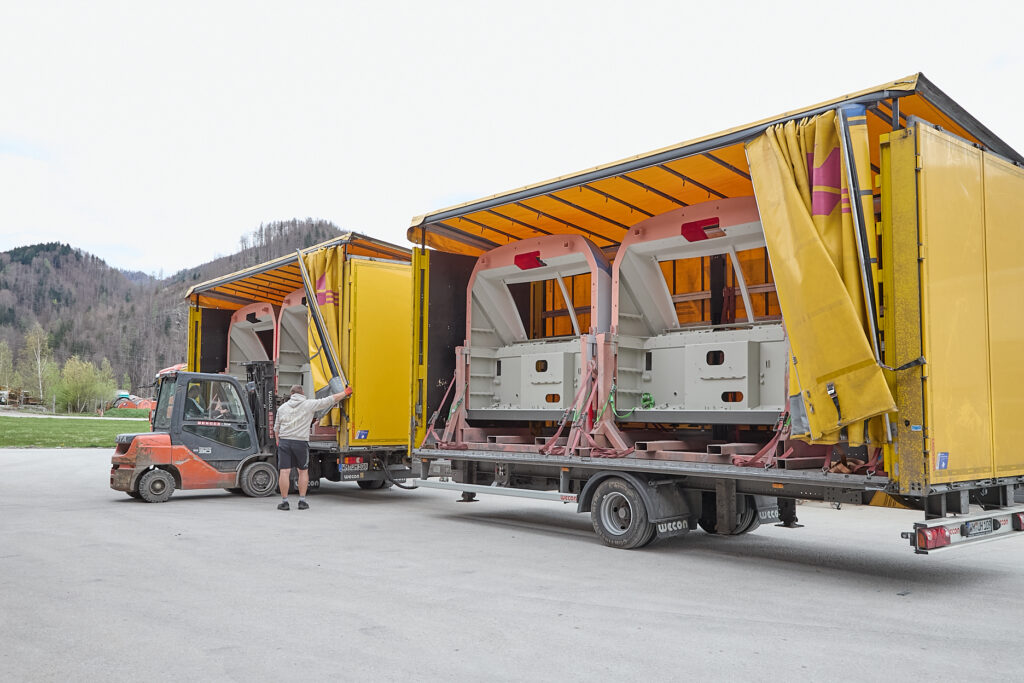 Ein Arbeiter benutzt einen Gabelstapler in der Nähe von zwei offenen Lastwagen, die mit großen Industriemaschinen beladen sind, mit Bäumen und Hügeln im Hintergrund. LAC Berger Industrielackierung
